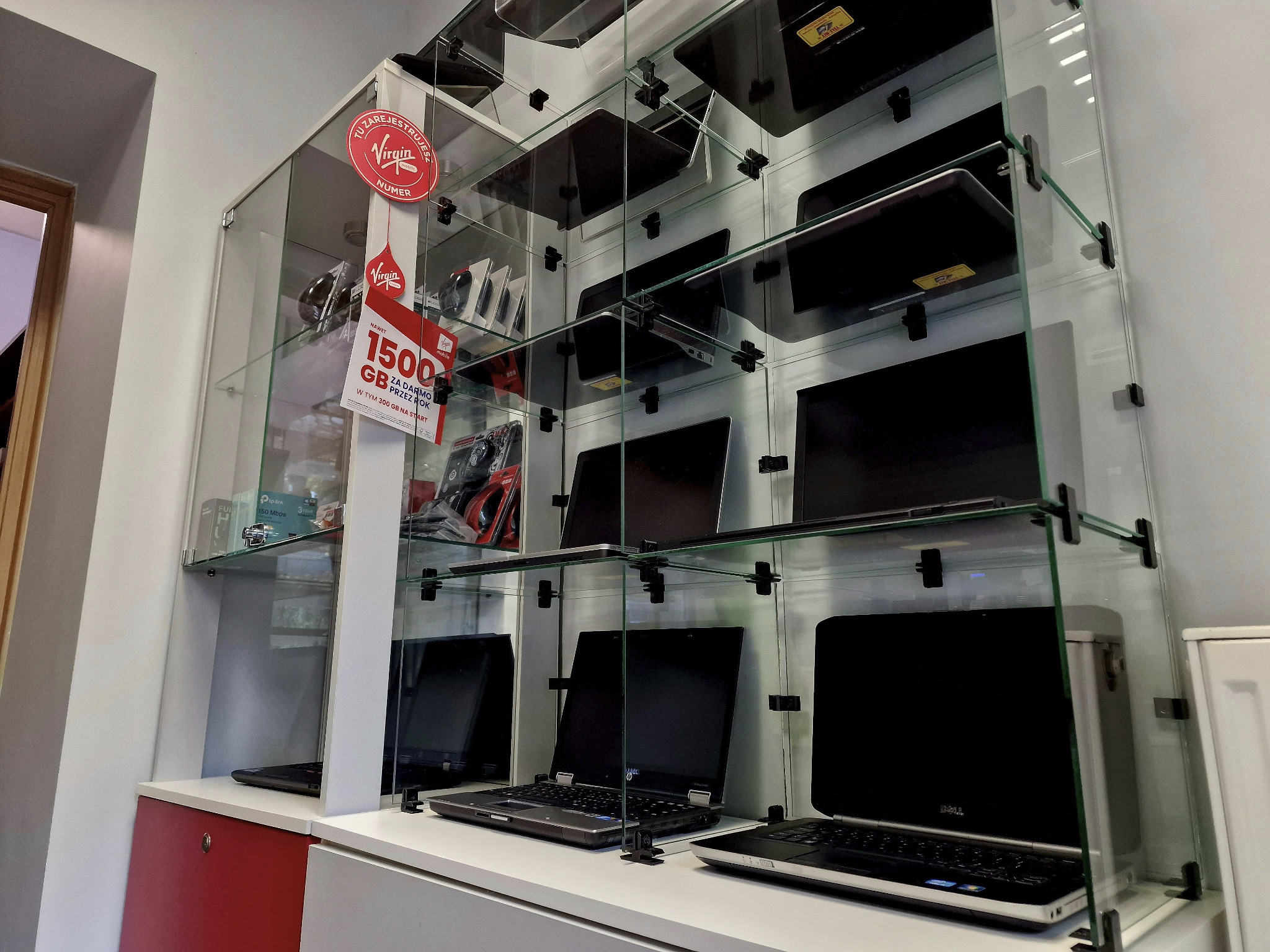 Wystawa laptopów różnych marek w szklanej gablocie w punkcie sprzedaży, widoczne logo operatora komórkowego z ofertą promocyjną na tle urządzeń.