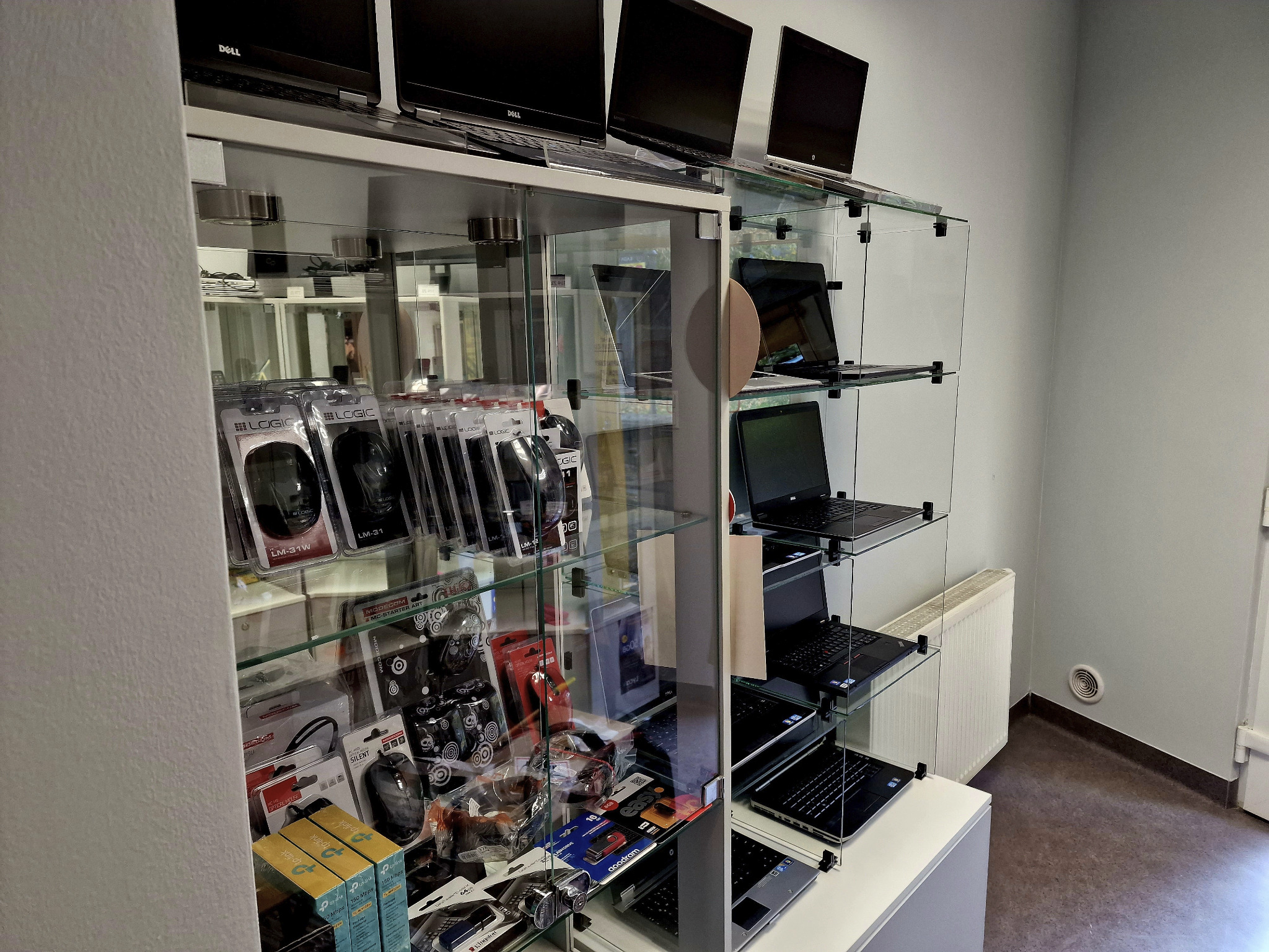 Szklana witryna sklepowa z laptopami Dell i akcesoriami komputerowymi, takimi jak myszki Logic i routery TP-Link, ustawionymi na półkach.