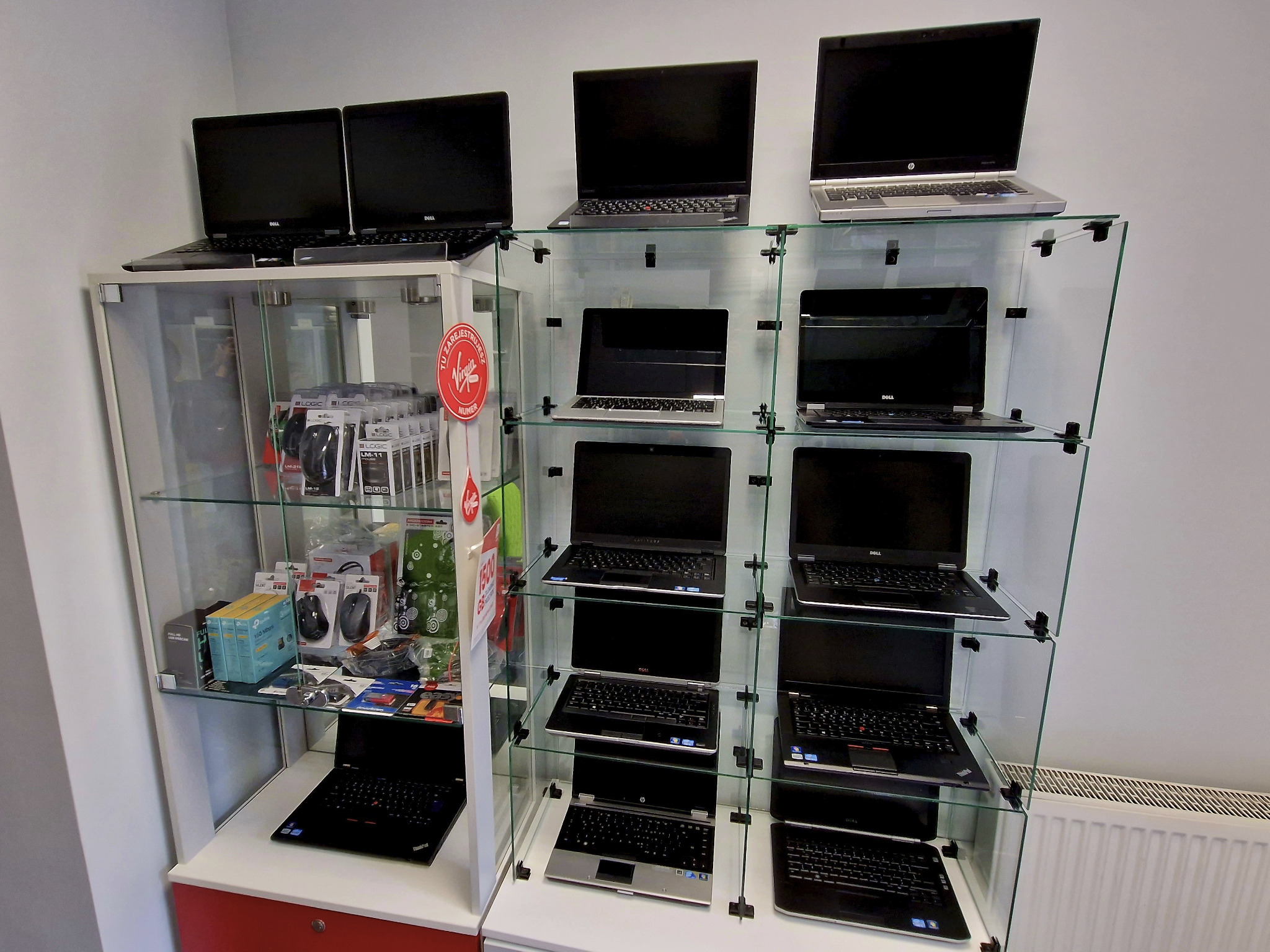 Szeroki kadr na szklaną witrynę z kilkunastoma używanymi laptopami różnych marek (Dell, HP, Lenovo) na półkach, obok myszy i akcesoria komputerowe w opakowaniach.