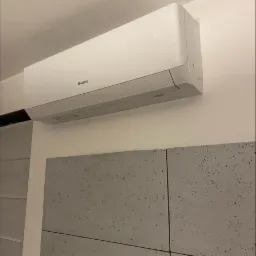 Biały klimatyzator marki Gree zamontowany na ścianie nad dekoracyjnymi panelami imitującymi beton.