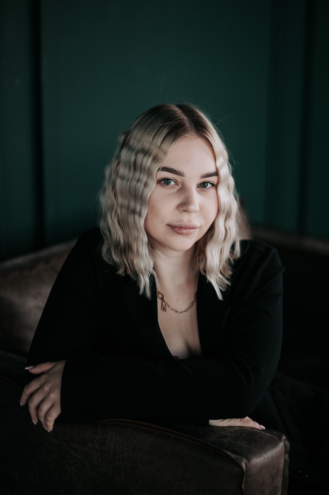 Portret kobiety w czarnej marynarce z falowanymi blond włosami i złotym naszyjnikiem z inicjałami, wykonany we Wrocławiu na tle ciemnozielonej ściany.