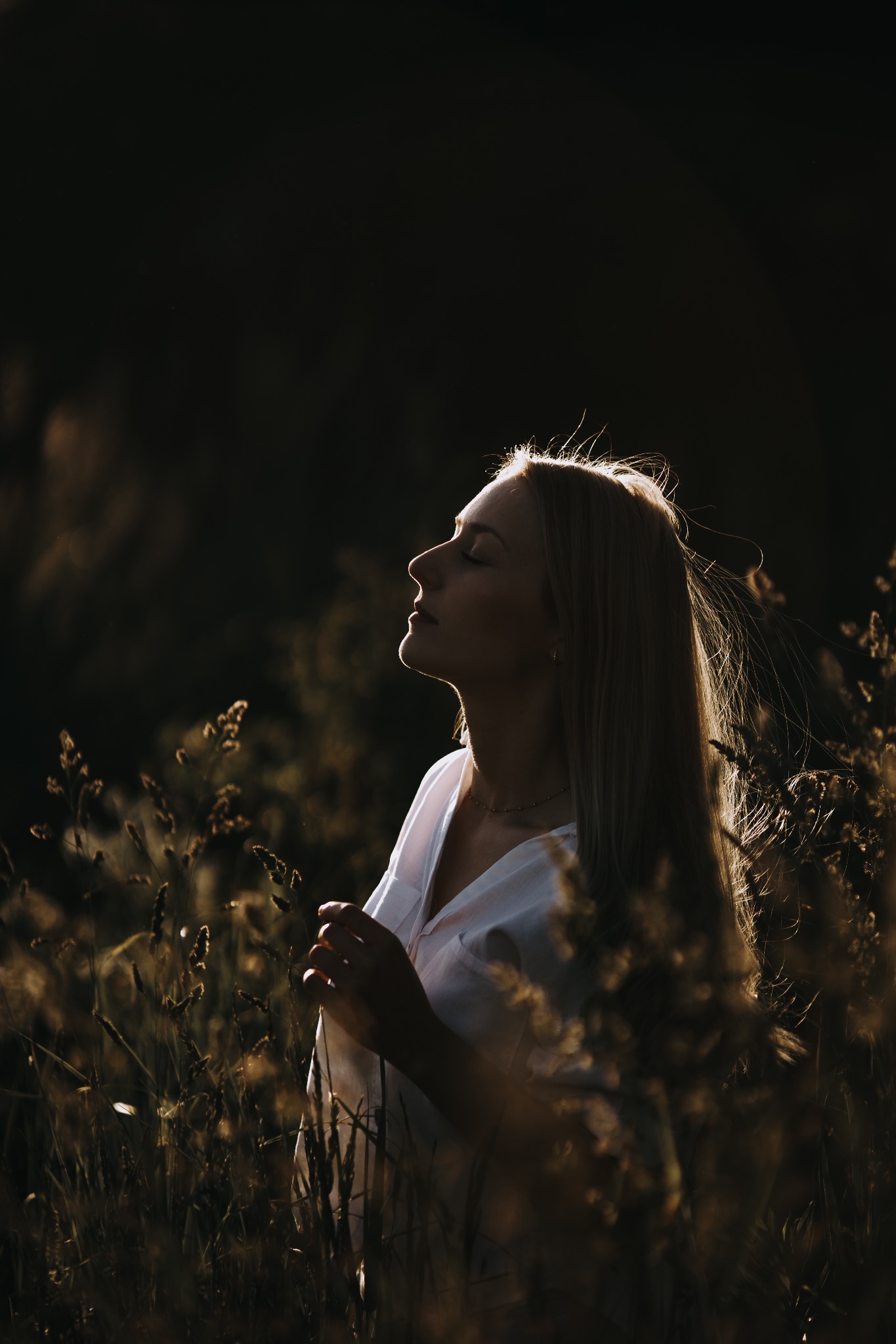 Portret kobiety z blond włosami, w białej koszuli, stojącej na tle wysokich traw w złotym świetle zachodzącego słońca, z zamkniętymi oczami.