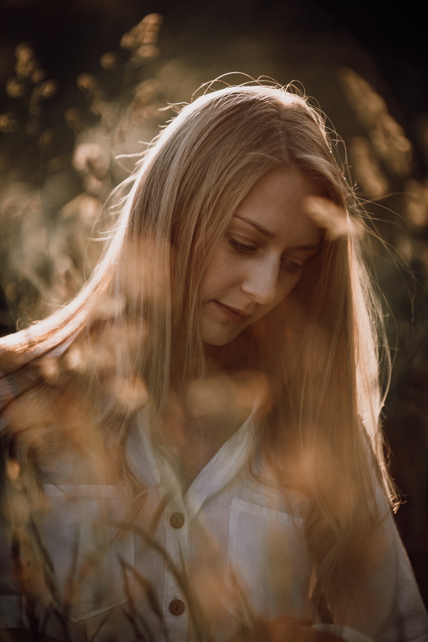 Portret młodej kobiety z długimi blond włosami, spuszczonym wzrokiem, w białej koszuli na tle rozmytej zieleni i złotych traw w kontraście ze światłem.