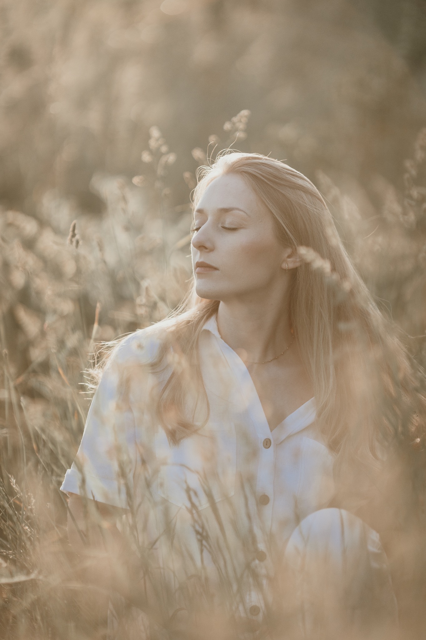 Portret kobiety z blond włosami, w białej koszuli, siedzącej pośród wysokich traw w miękkim, rozproszonym świetle, z zamkniętymi oczami.
