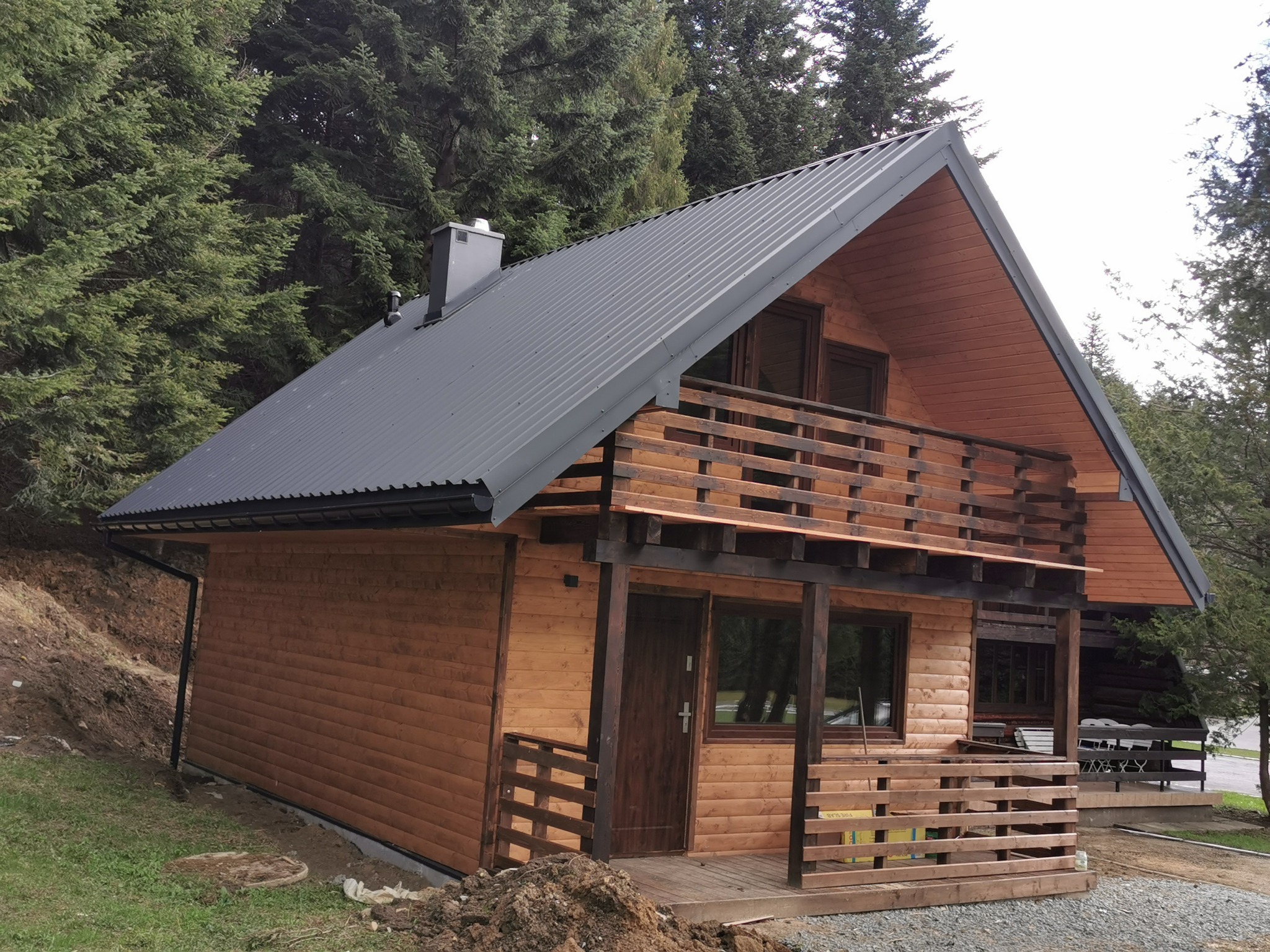 Gotowy drewniany dom z szarą blachodachówką, balkonem z drewnianą balustradą i widocznymi elementami konstrukcyjnymi, usytuowany na tle gęstego lasu iglastego.