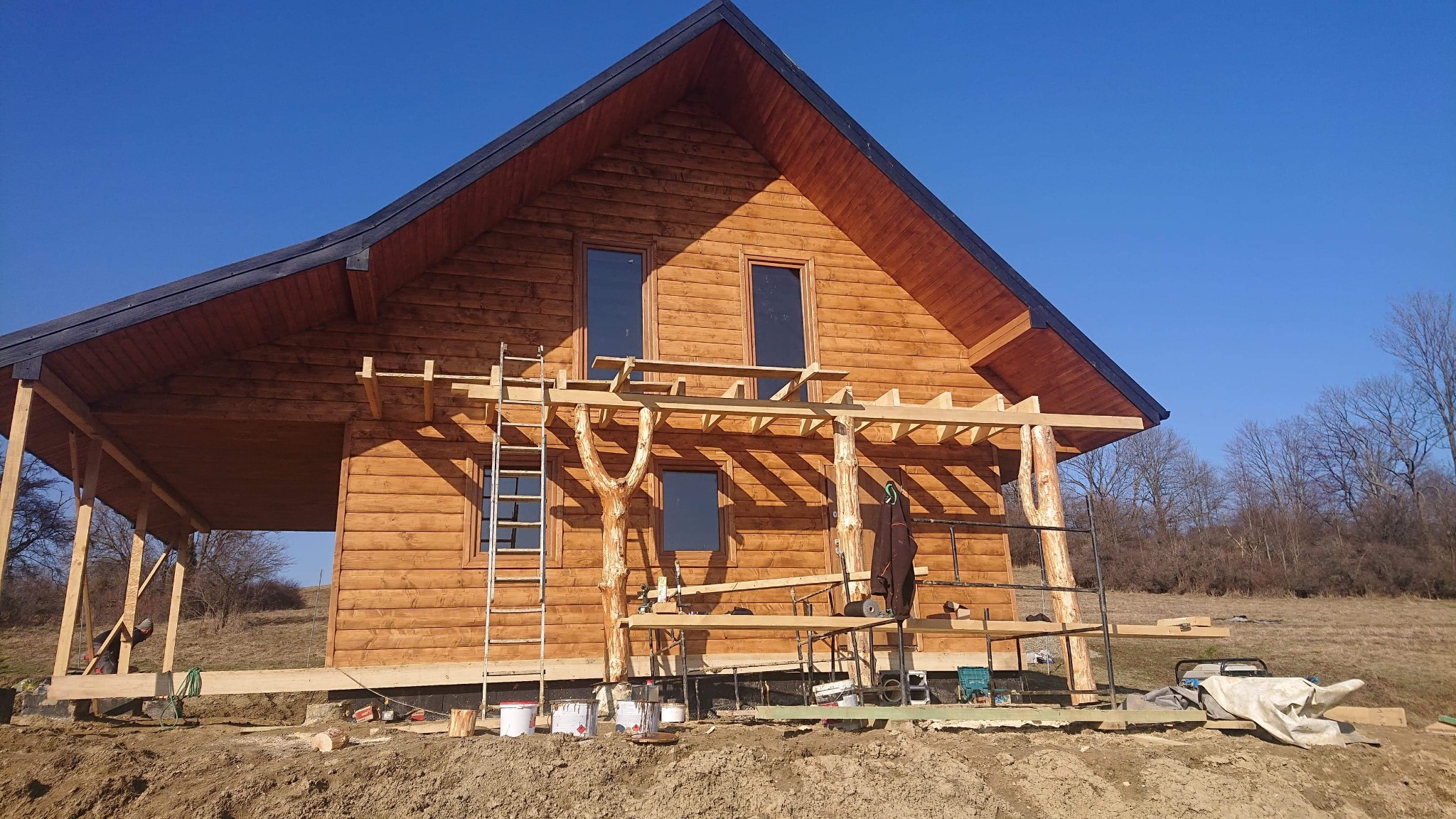 Surowy, drewniany dom w trakcie budowy, z widocznym rusztowaniem i elementami konstrukcyjnymi werandy, na tle błękitnego nieba i jesiennego krajobrazu.