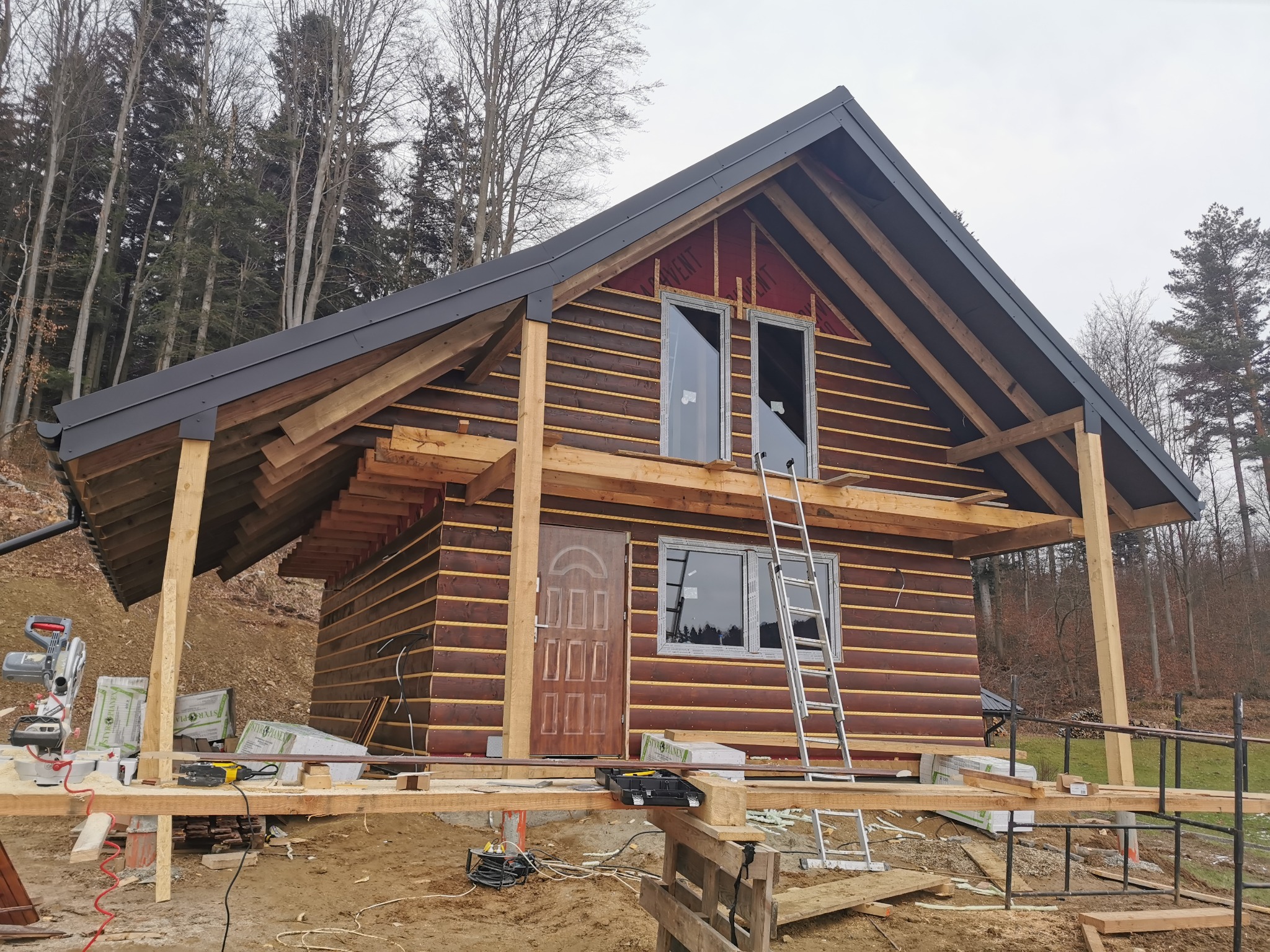 Dom szkieletowy w trakcie budowy z widoczną konstrukcją drewnianą, oknami bez szyb, drabina oparta o ścianę, narzędzia budowlane na rusztowaniu i las w tle.