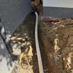 Biała perforowana rura drenażowa ułożona w wykopie wzdłuż fundamentu budynku z elewacją w odcieniach szarości, zasypana częściowo żwirem.