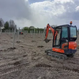 Pomarańczowa mini koparka Kubota podnosi betonowy słup na polu z rzędami ustawionych słupów, w tle widoczny traktor.