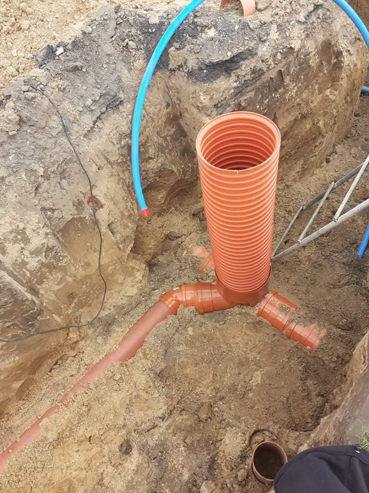 Pomarańczowe rury drenażowe złączone w wykopie, widoczne niebieskie rury i fragment drabiny.