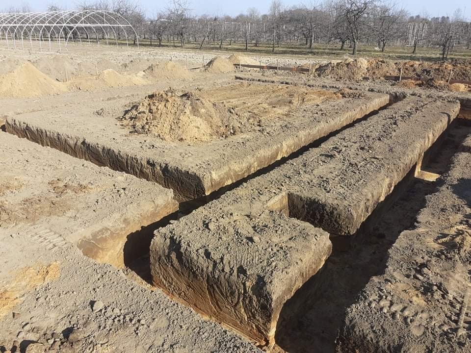 Wykonane wykopy pod fundamenty domu jednorodzinnego, widoczne ślady opon na ziemi, w tle szklarnia tunelowa.