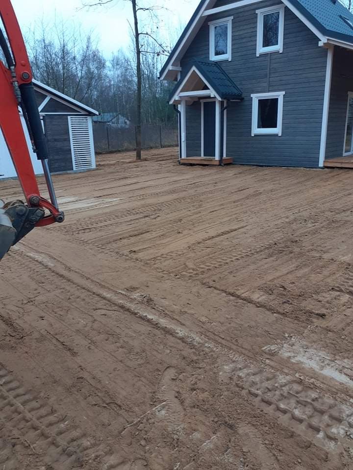 Nierówny teren z widocznymi śladami opon po maszynach budowlanych przed nowo wybudowanym domem z szarą elewacją i białymi obramowaniami okien i drzwi, w tle widoczny mały budynek gospodarczy i drzewa.