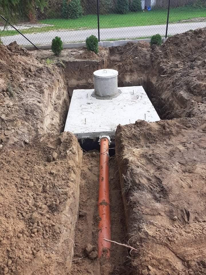 Betonowy zbiornik z pokrywą umieszczony w wykopie, z podłączoną rurą kanalizacyjną, otoczony ziemią i zielenią w tle.