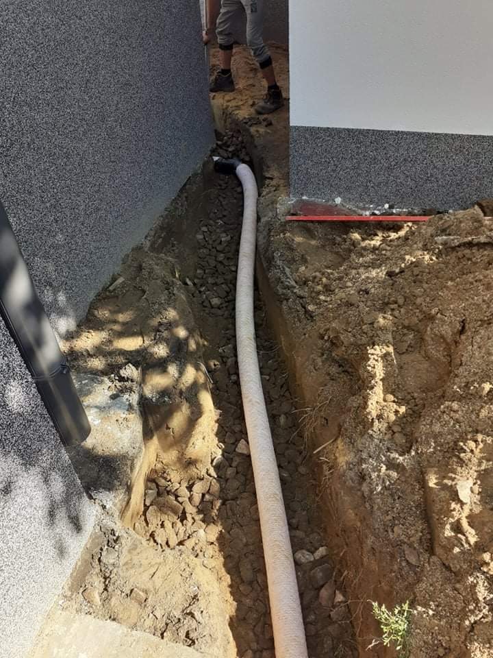 Biała perforowana rura drenażowa ułożona w wykopie wzdłuż fundamentu budynku z elewacją w odcieniach szarości, zasypana częściowo żwirem.