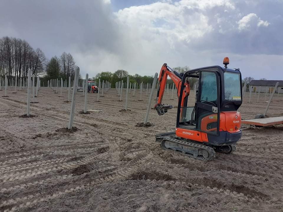 Pomarańczowa mini koparka Kubota podnosi betonowy słup na polu z rzędami ustawionych słupów, w tle widoczny traktor.