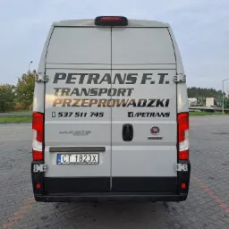 Tył szarego busa Fiat Ducato Maxi firmy PETRANS F.T. z numerem telefonu i informacją o transporcie i przeprowadzkach, zaparkowanego na brukowanym placu.