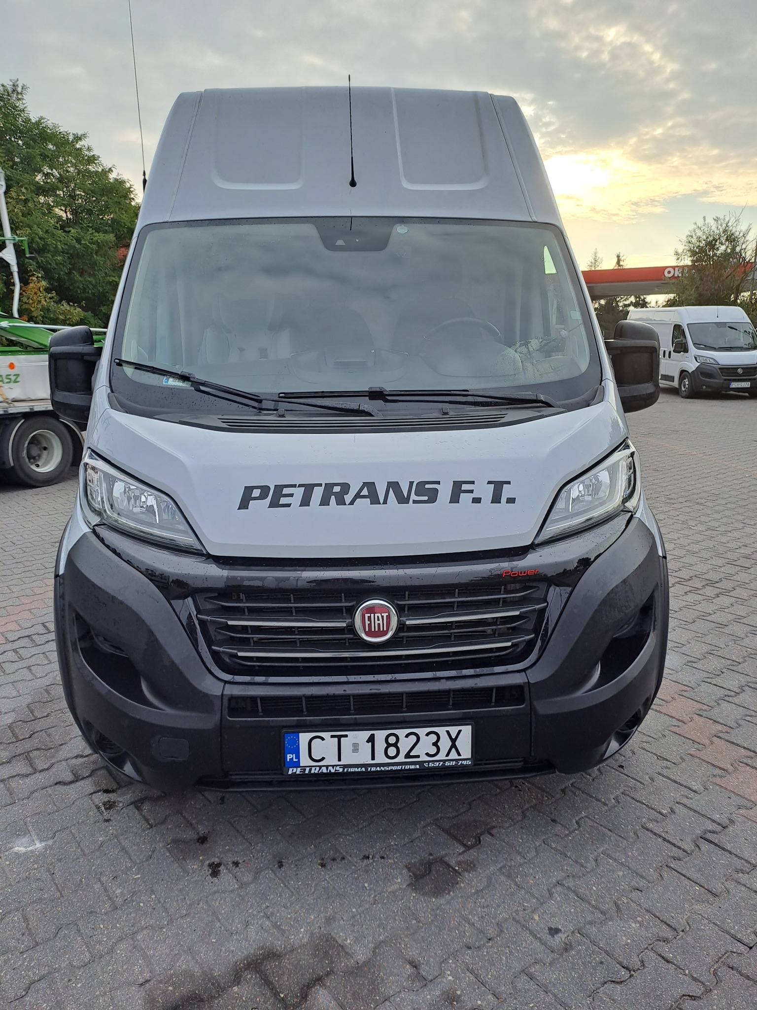 Szary bus marki Fiat z napisem PETRANS F.T. na tle stacji benzynowej i zachmurzonego nieba.