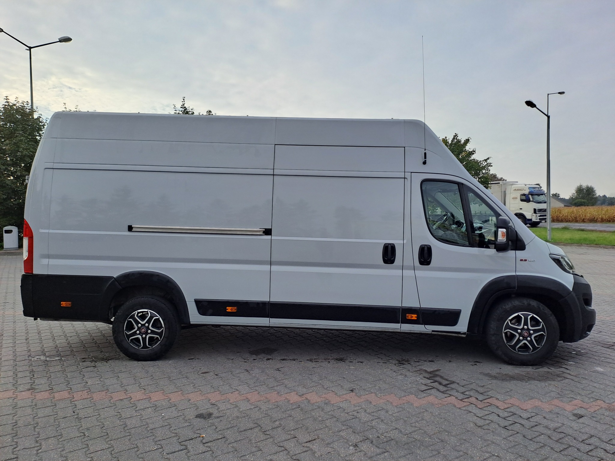 Szary, wysoki bus dostawczy zaparkowany na brukowanym parkingu, widok z boku, z widocznymi detalami opon i felg.