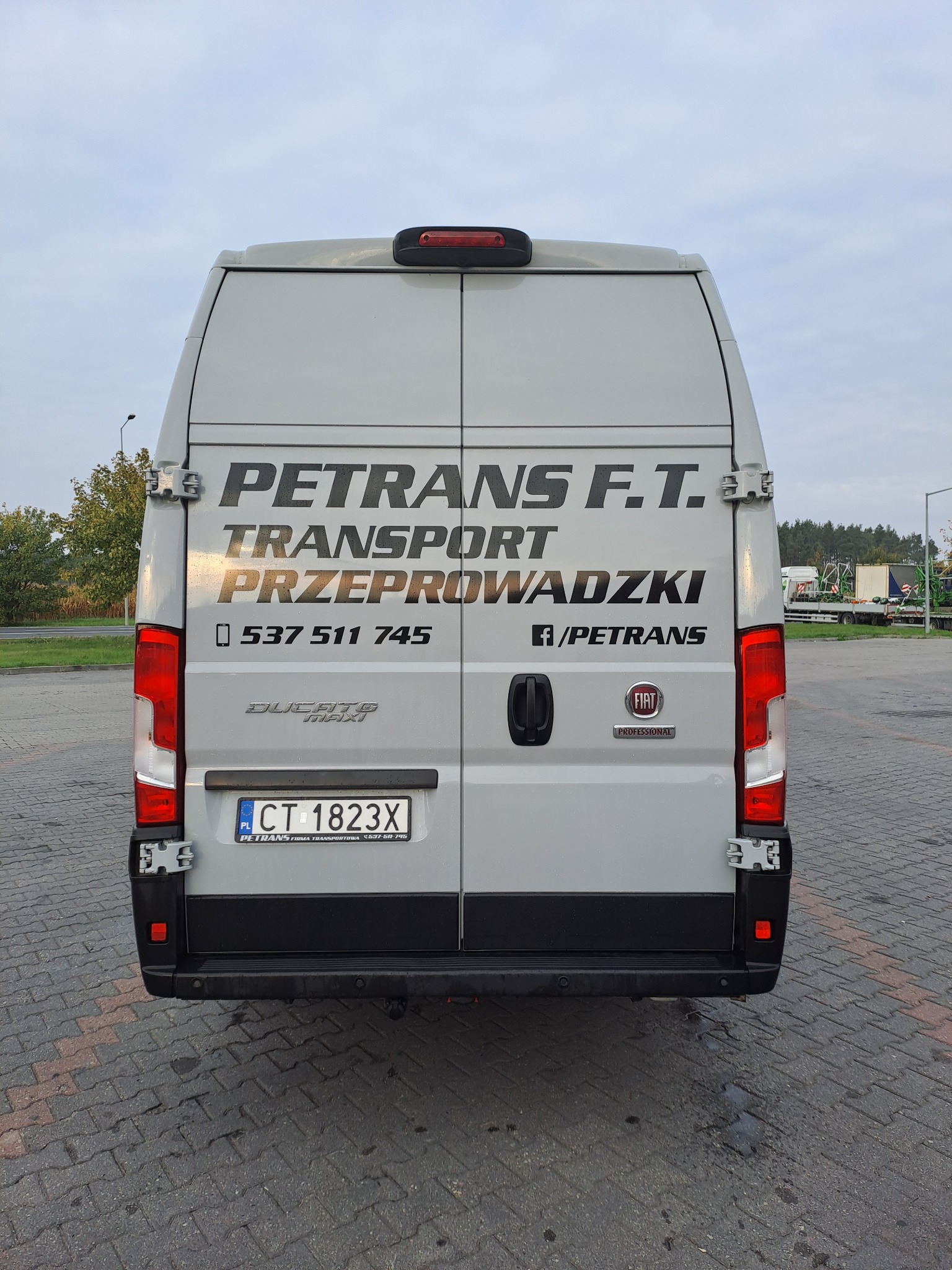 Tył szarego busa Fiat Ducato Maxi firmy PETRANS F.T. z numerem telefonu i informacją o transporcie i przeprowadzkach, zaparkowanego na brukowanym placu.