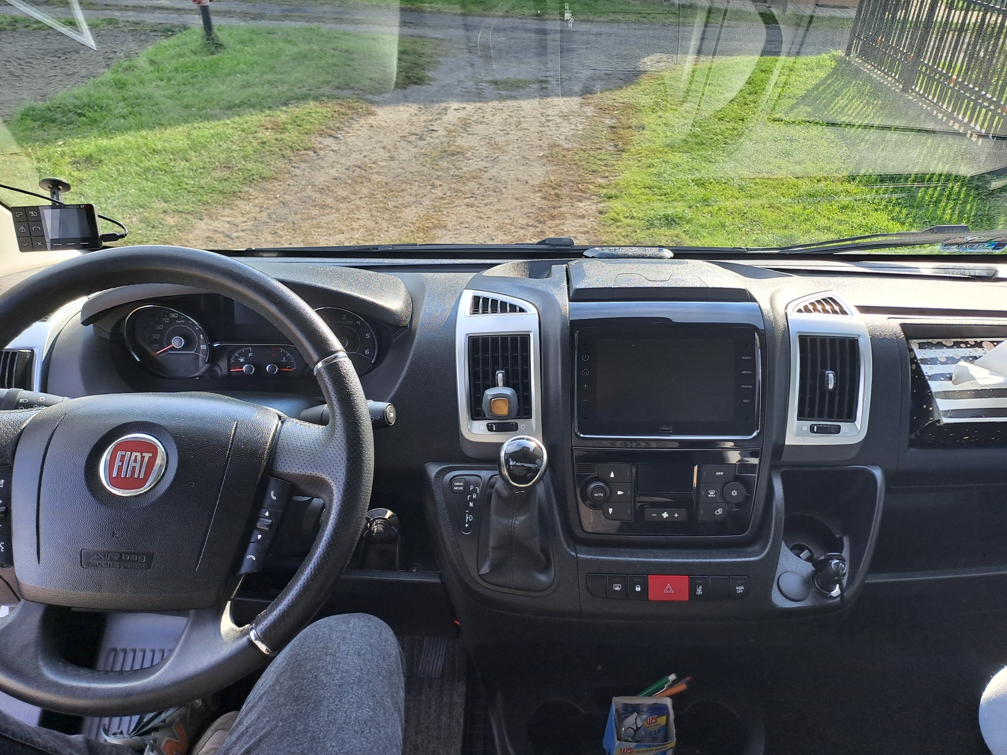 Wnętrze kabiny Fiata Ducato z widokiem na deskę rozdzielczą, kierownicę z logo FIAT i nawigację samochodową podczas jazdy po żwirowej drodze.