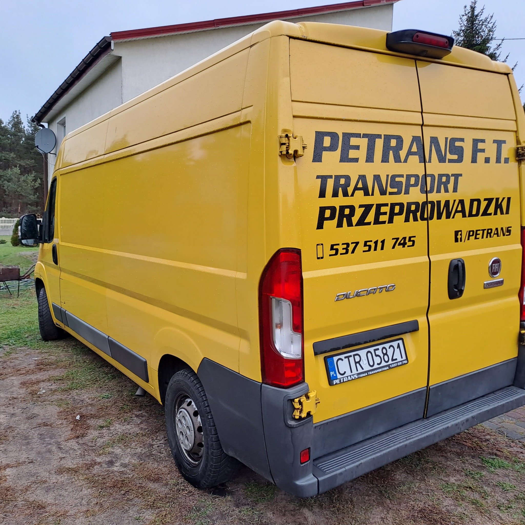 Żółty bus marki Fiat Ducato z czarnym napisem 'PETRANS F.T. TRANSPORT PRZEPROWADZKI 537 511 745 f/PETRANS' na tylnych drzwiach, zaparkowany na zewnątrz budynku w pochmurny dzień.