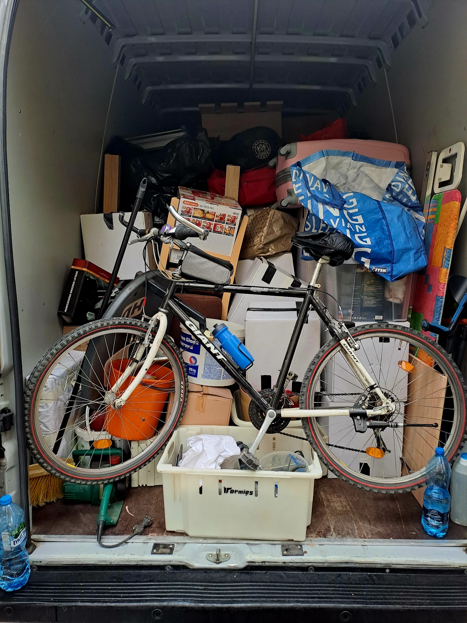 Wnętrze busa wypełnione różnorodnymi przedmiotami, w tym rower Giant na pierwszym planie, pudełka, torby i narzędzia, tworzące wrażenie załadunku przed transportem.