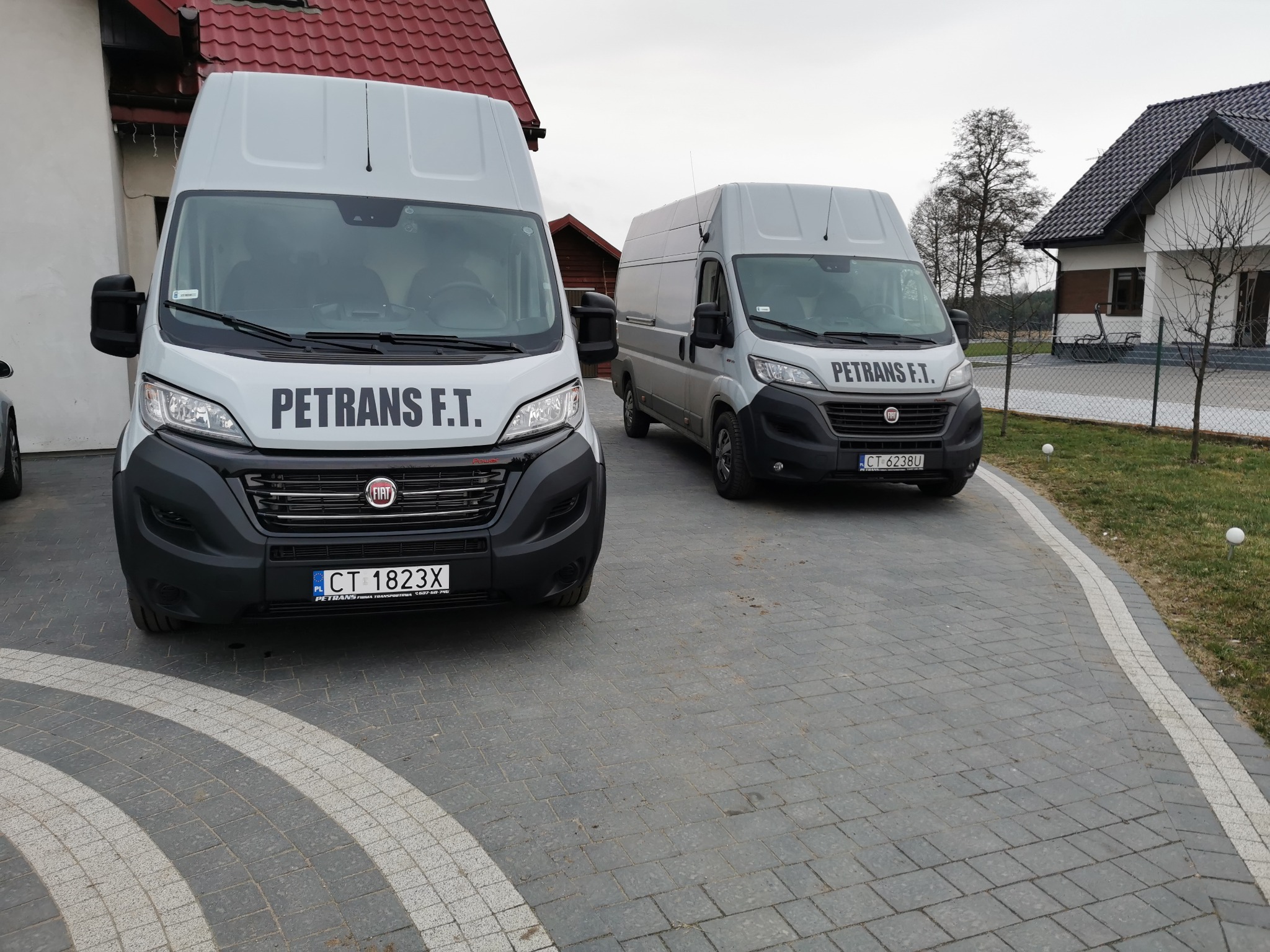 Dwa busy firmy PETRANS F.T. zaparkowane na brukowanym podjeździe przed domami z czerwonymi dachami.