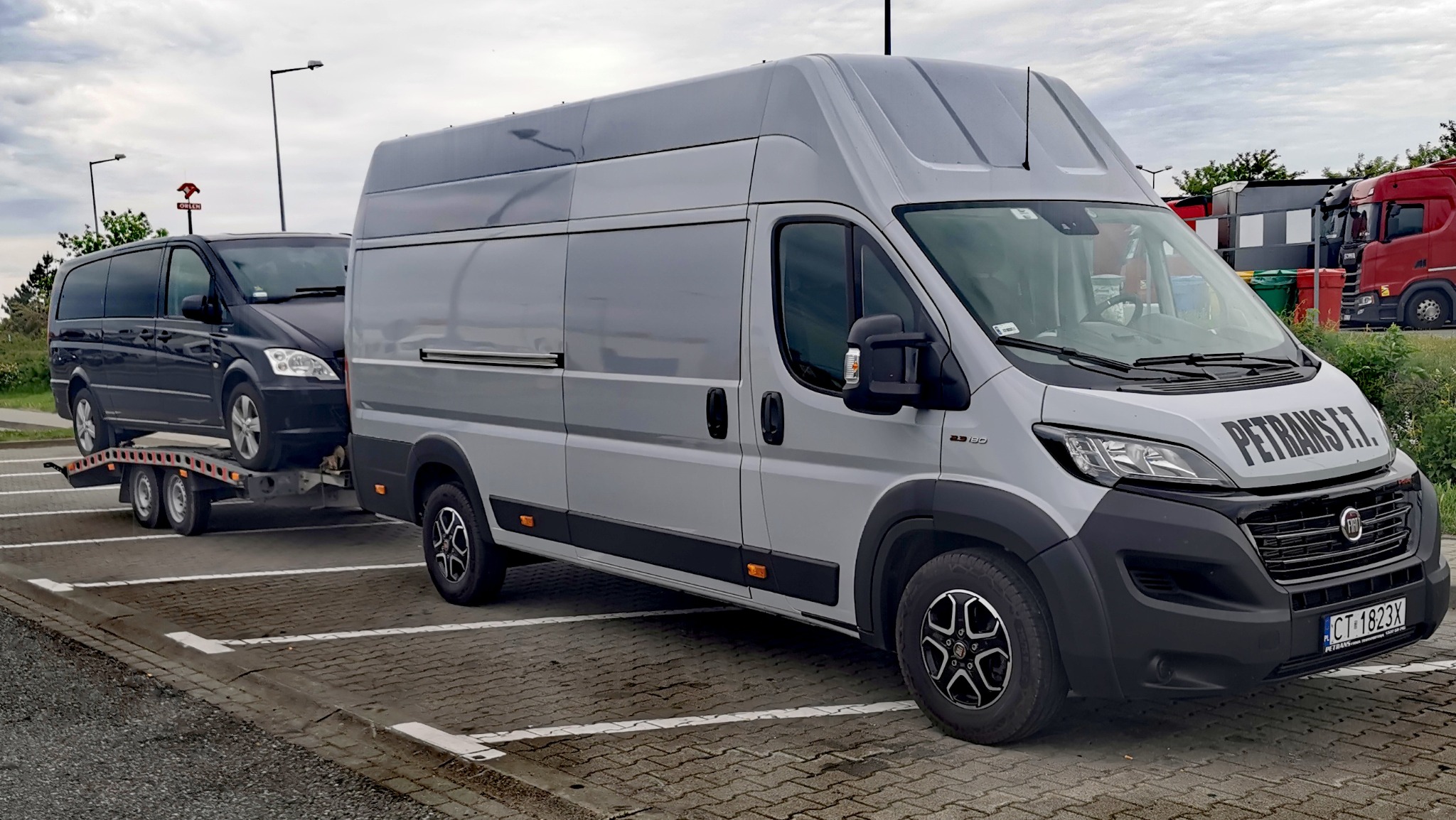Szary bus Fiat Ducato z napisem PETRANS F.T. ciągnie przyczepę z czarnym vanem Mercedes na parkingu.