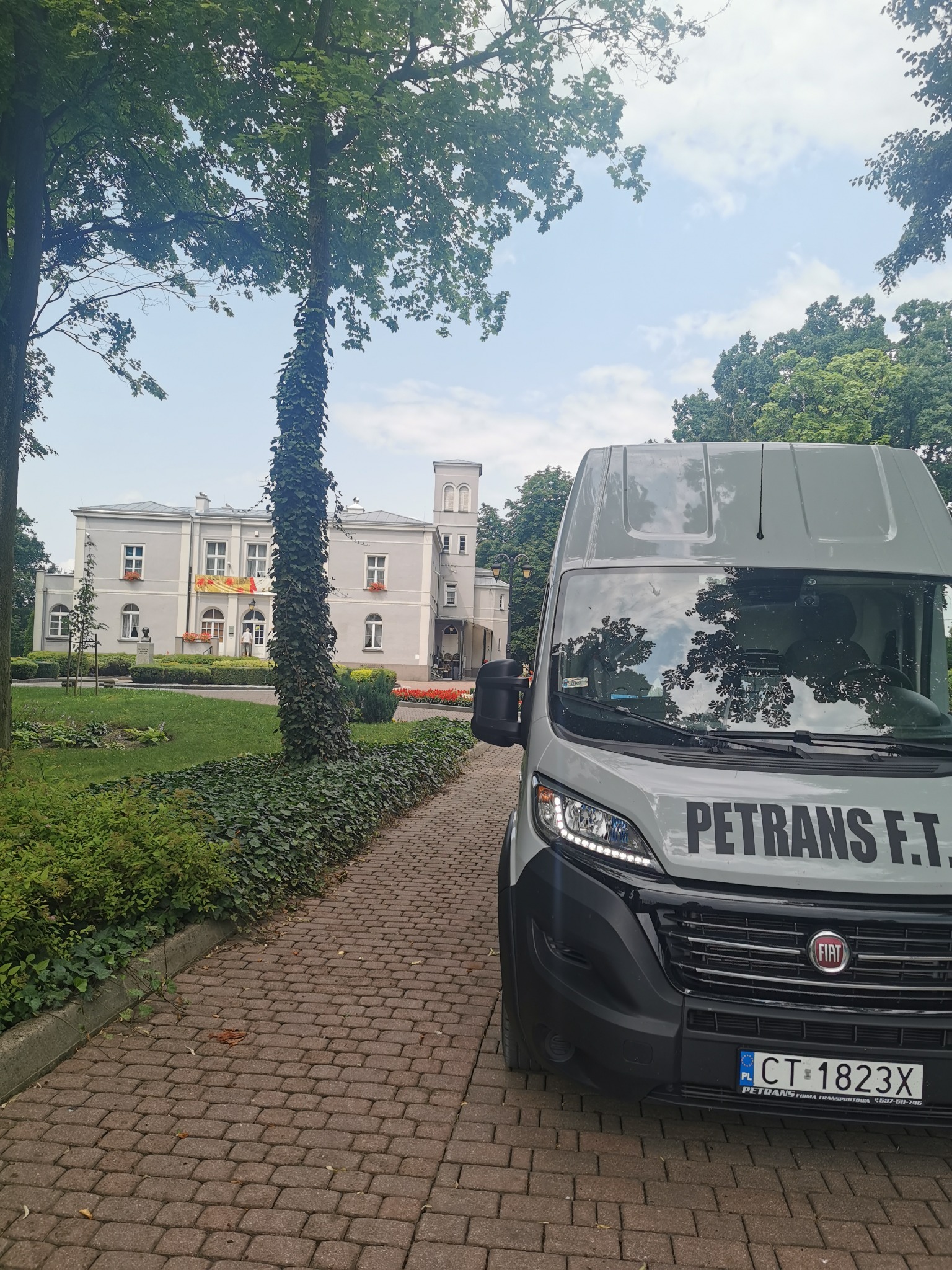Szary bus firmy PETRANS F.T. zaparkowany na brukowanej ścieżce przed budynkiem w otoczeniu zieleni.