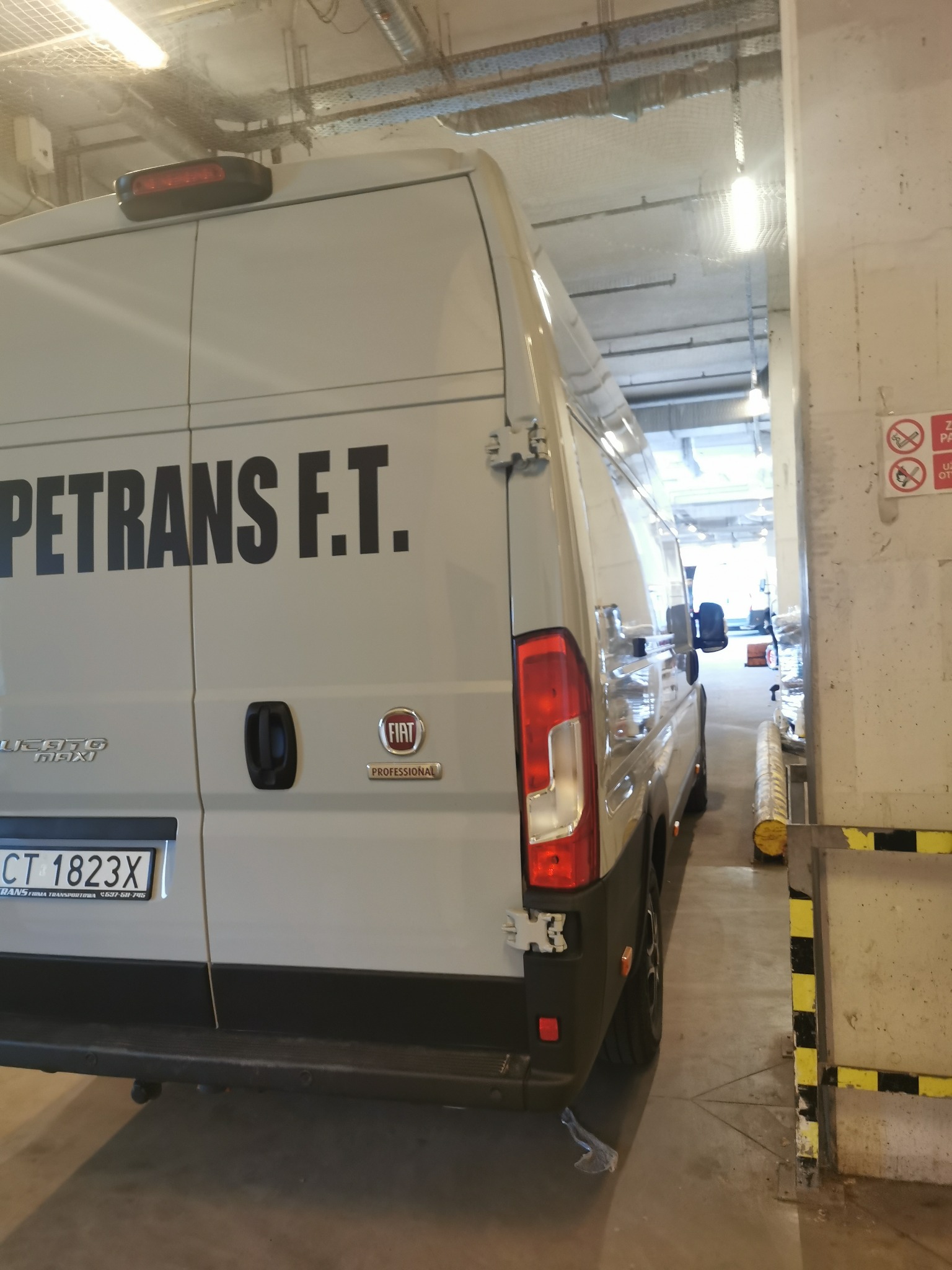 Biały bus marki Fiat Ducato z napisem PETRANS F.T. zaparkowany w hali garażowej, widok z tyłu i boku, z widocznymi oznaczeniami firmy transportowej.