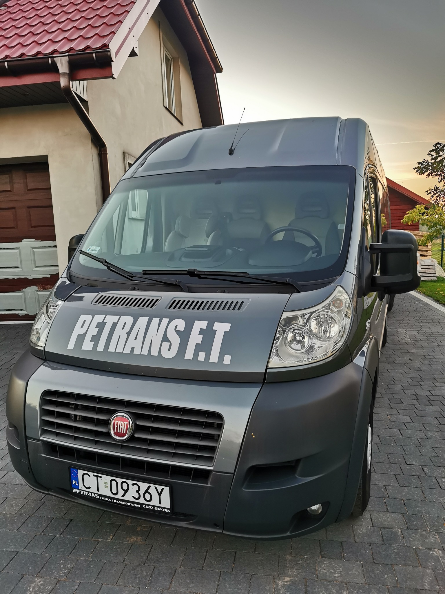Szary bus firmy PETRANS F.T. zaparkowany na brukowanym podjeździe przed domem z czerwonym dachem.