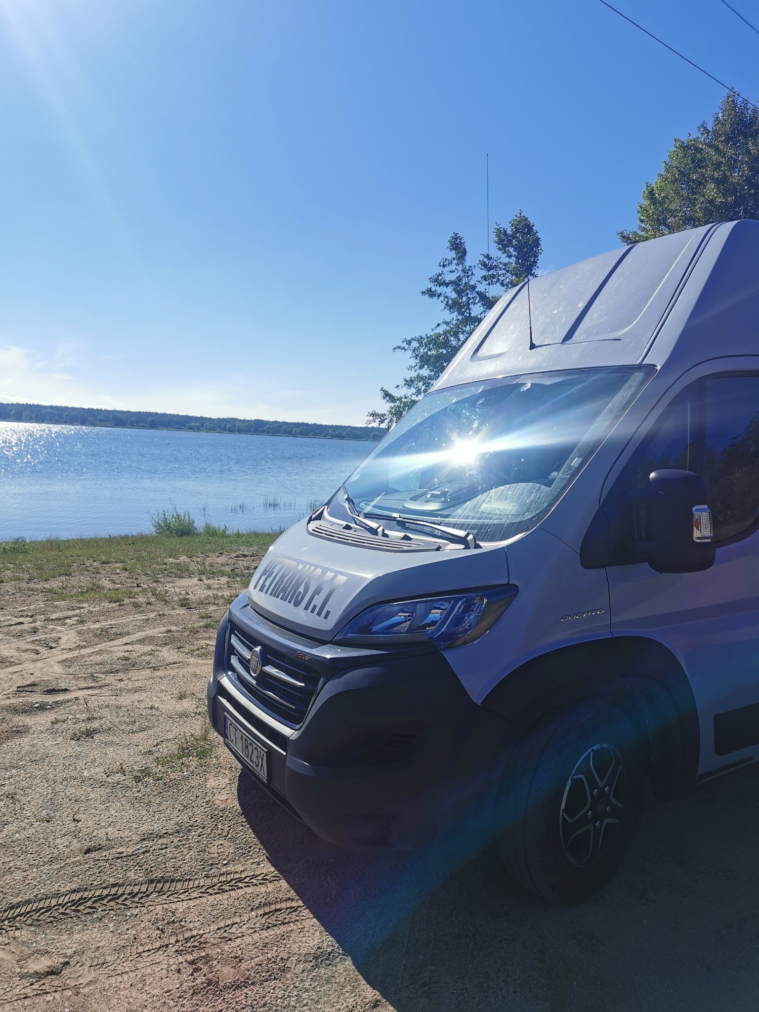 Jasnoszary bus marki Fiat z napisem 'PETRANS S.I.' zaparkowany na piaszczystym brzegu jeziora w słoneczny dzień.