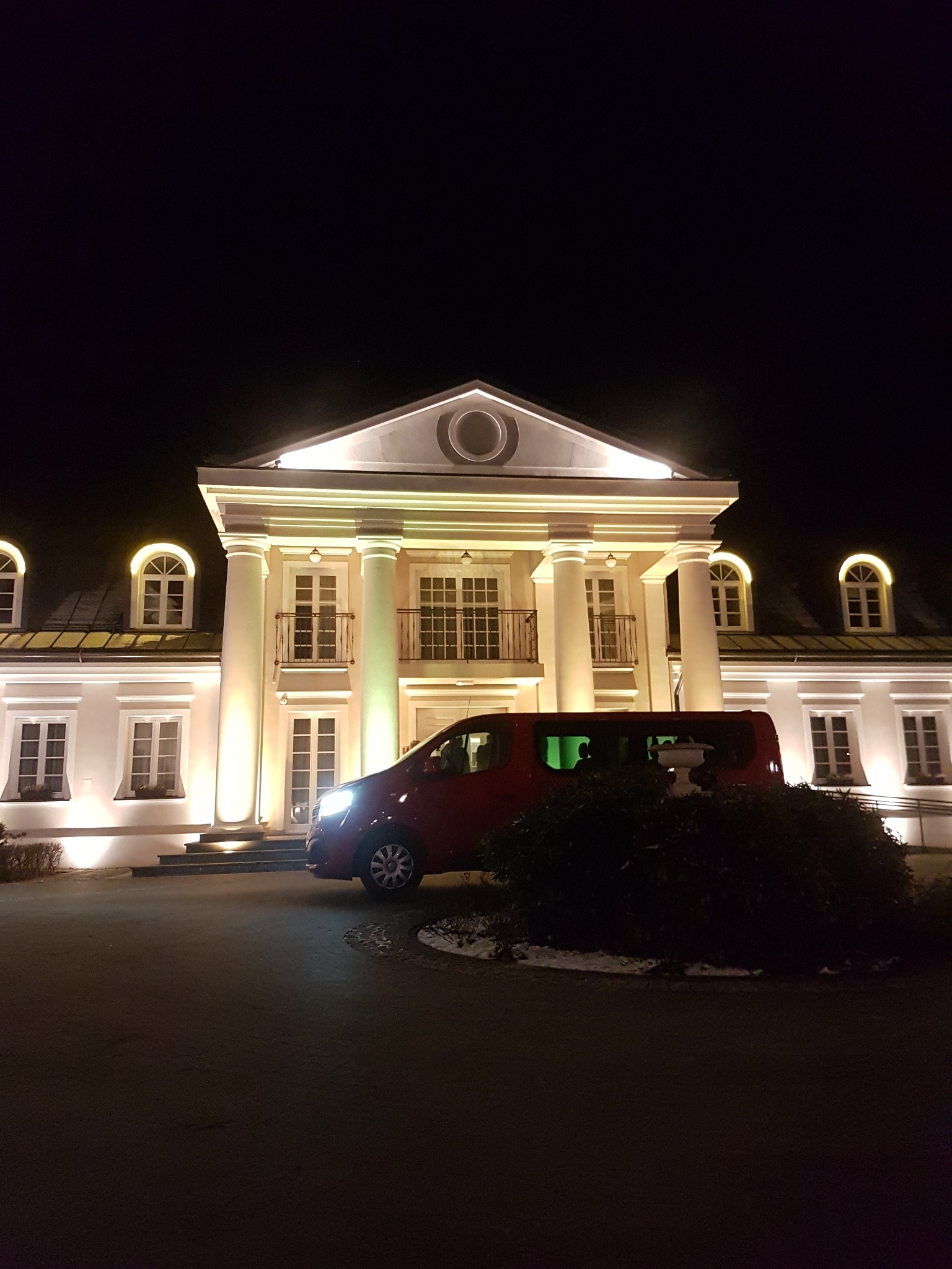 Czerwony van na tle podświetlonego, klasycystycznego budynku z kolumnami, nocne ujęcie.