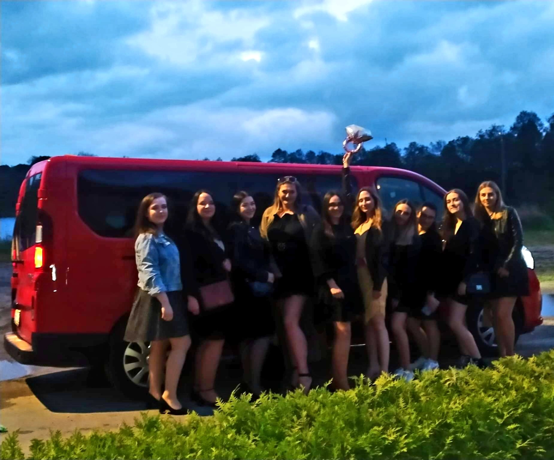 Grupa kobiet świętuje przed czerwonym vanem w Rawa Mazowiecka, jedna z nich trzyma balon w kształcie pierścionka zaręczynowego, sugerując wieczór panieński.