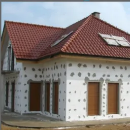 Dom jednorodzinny w trakcie ocieplania, widoczne płyty styropianowe z przyklejonymi plackami zaprawy, przygotowany do dalszych prac elewacyjnych. Dach pokryty czerwoną dachówką.