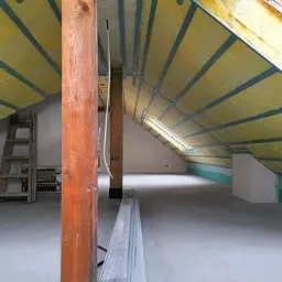 Poddasze w trakcie ocieplania: żółta wełna mineralna między krokwiami, widoczne okno dachowe, drewniany słup konstrukcyjny i metalowe profile na podłodze.