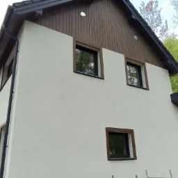 Wykończona elewacja domu z jasnym tynkiem barankiem, brązowymi ramami okiennymi i drewnianym wykończeniem pod dachem.