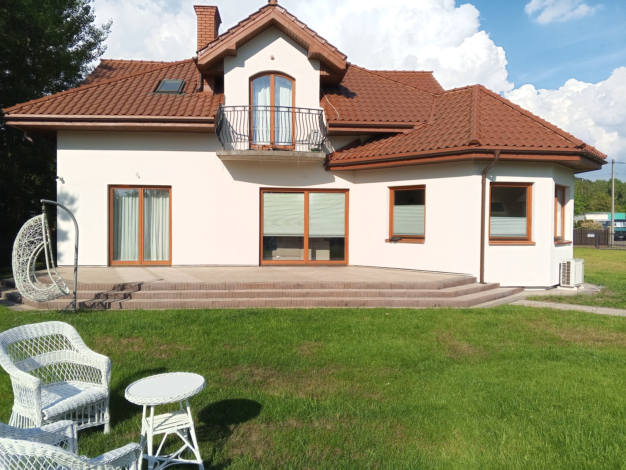 Biały dom jednorodzinny z brązowym dachem z dachówki ceramicznej, balkonem i schodami z kostki brukowej, otoczony zielonym trawnikiem i białymi meblami ogrodowymi.