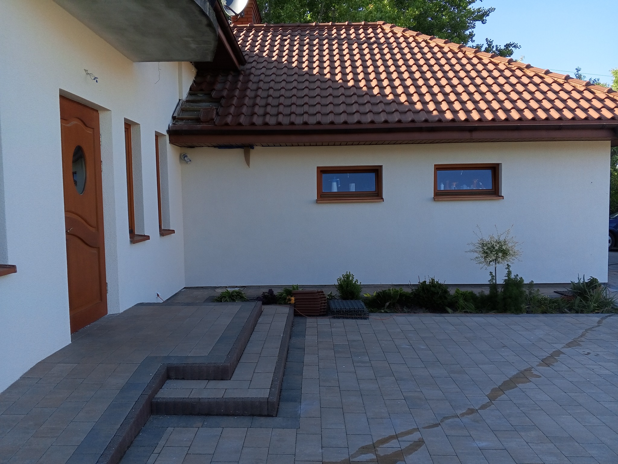 Biała elewacja domu jednorodzinnego z brązową dachówką, widoczne dwa okna i brązowe drzwi wejściowe, teren wokół domu wyłożony kostką brukową, widoczne rośliny ozdobne.