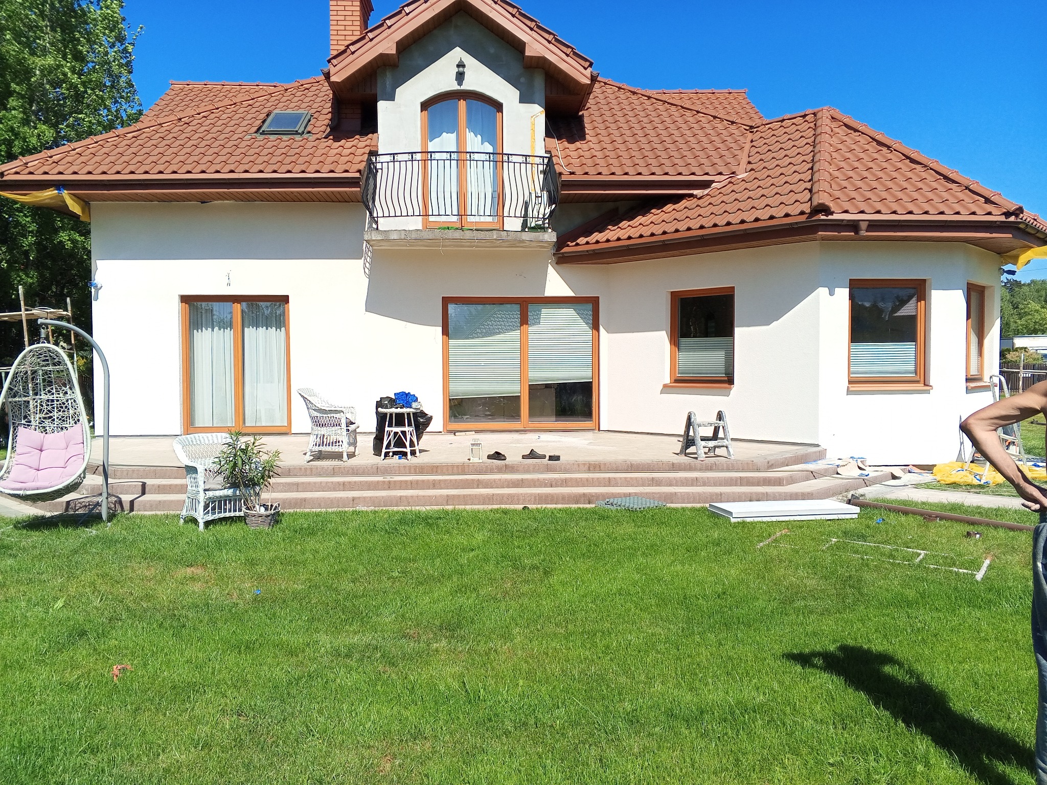 Jasna elewacja domu jednorodzinnego z brązową dachówką, widoczne schody prowadzące do drzwi tarasowych, krzesła wiklinowe i huśtawka wisząca, przygotowania do prac wykończeniowych na trawniku.