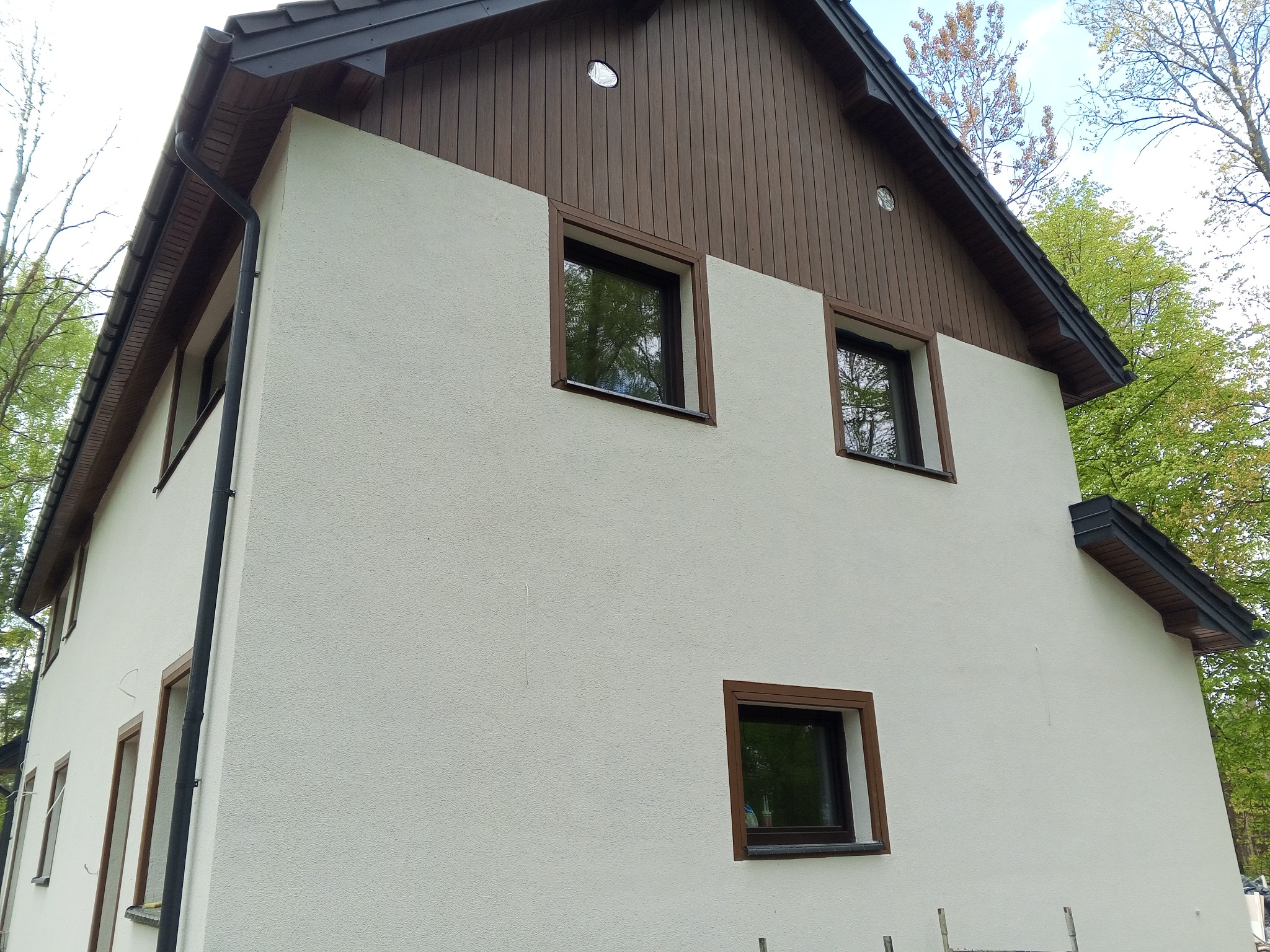 Wykończona elewacja domu z jasnym tynkiem barankiem, brązowymi ramami okiennymi i drewnianym wykończeniem pod dachem.