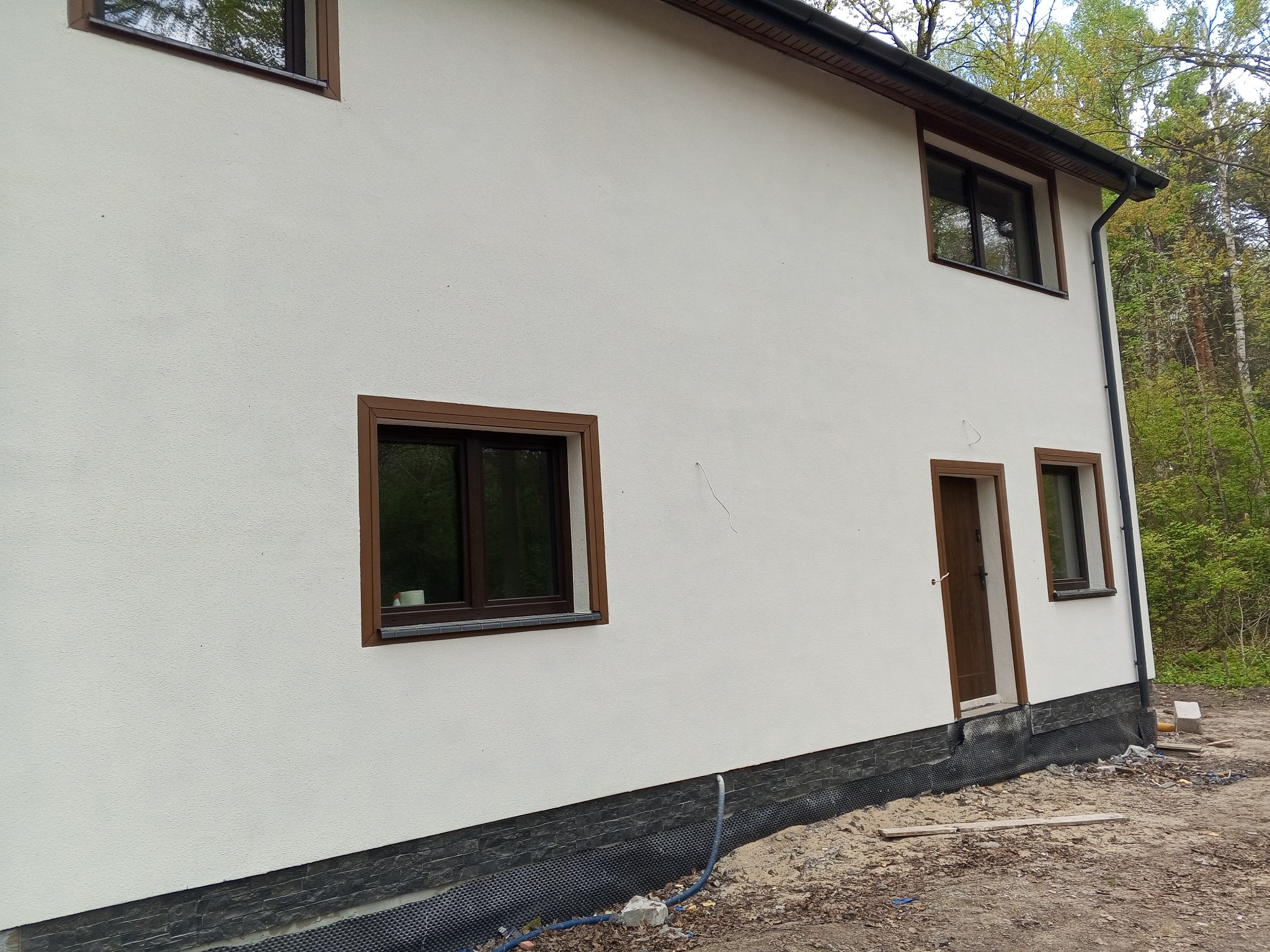 Boczny widok domu z jasną elewacją, brązowymi ramami okiennymi i drzwiami, wykończony cokołem z ciemnej cegły, otoczony drzewami.