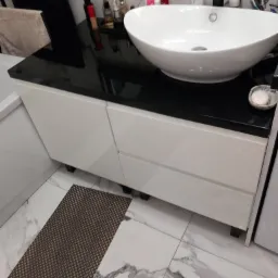 Biała umywalka nablatowa na szafce z białymi frontami i czarnym blatem, na marmurowej podłodze z brązowym dywanikiem łazienkowym.