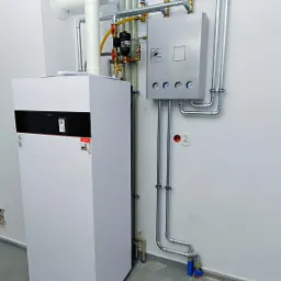 Nowoczesny, biały piec grzewczy z podłączonymi rurami miedzianymi i stalowymi, zbiornik wyrównawczy w kolorze niebieskim, szara szafka sterownicza z manometrami zamontowana na ścianie, widoczne...