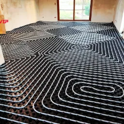 Białe rury ogrzewania podłogowego ułożone na czarnej macie w surowym pomieszczeniu z oknem i logo firmy HydroVIP w lewym górnym rogu.