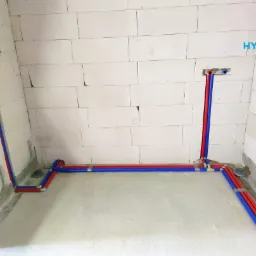 Nowa instalacja wodna z rur PEX, kolory niebieski i czerwony, prowadzone po ścianach z białego betonu komórkowego i wylewce podłogowej, widoczne złącza i logo firmy w prawym górnym rogu.