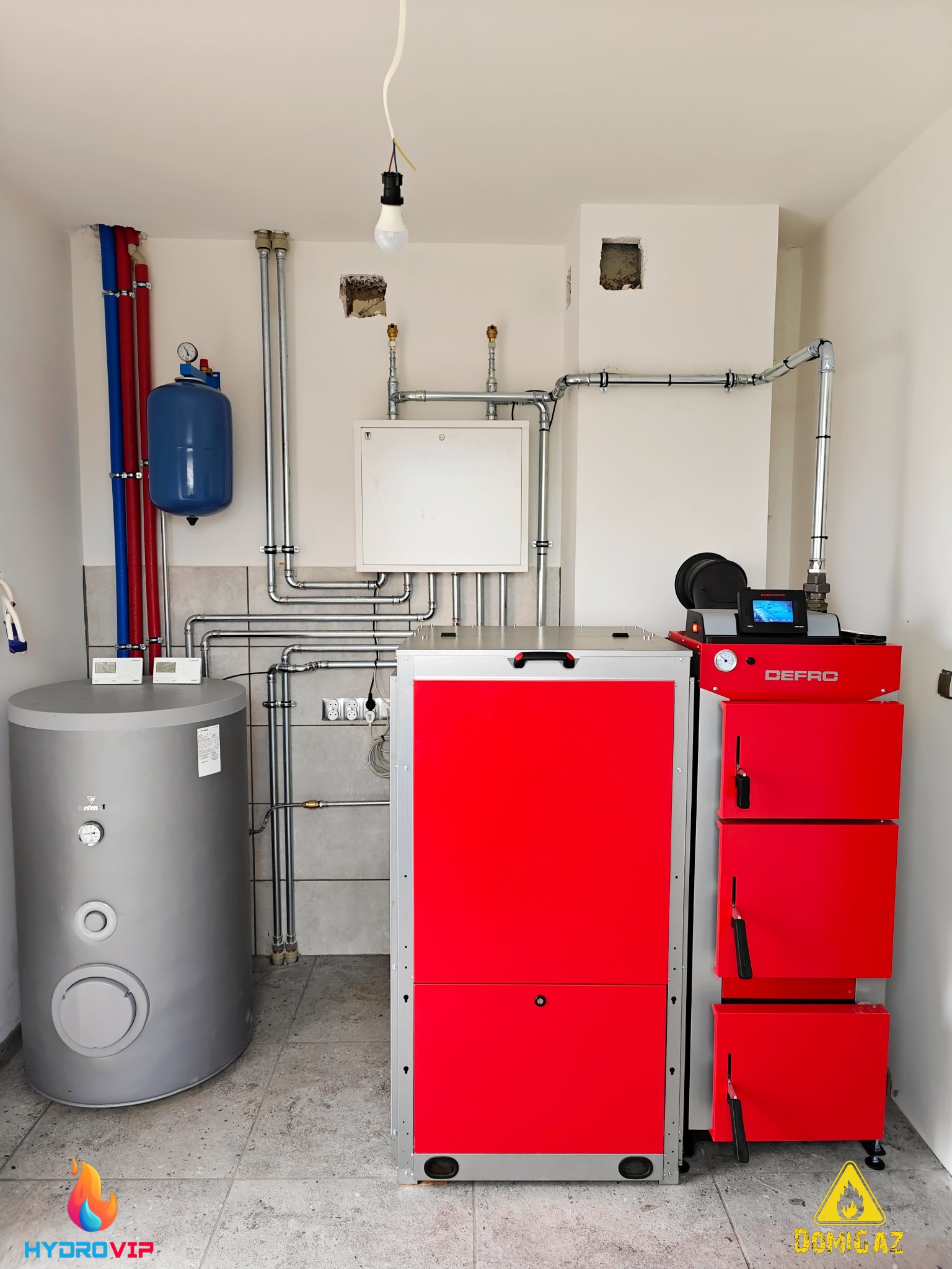 Kotłownia z piecem na paliwo stałe DEFRO w kolorze czerwonym, zasobnikiem wody w kolorze szarym, instalacją hydrauliczną z rurami w kolorze niebieskim i czerwonym oraz instalacją z rur stalowych,...