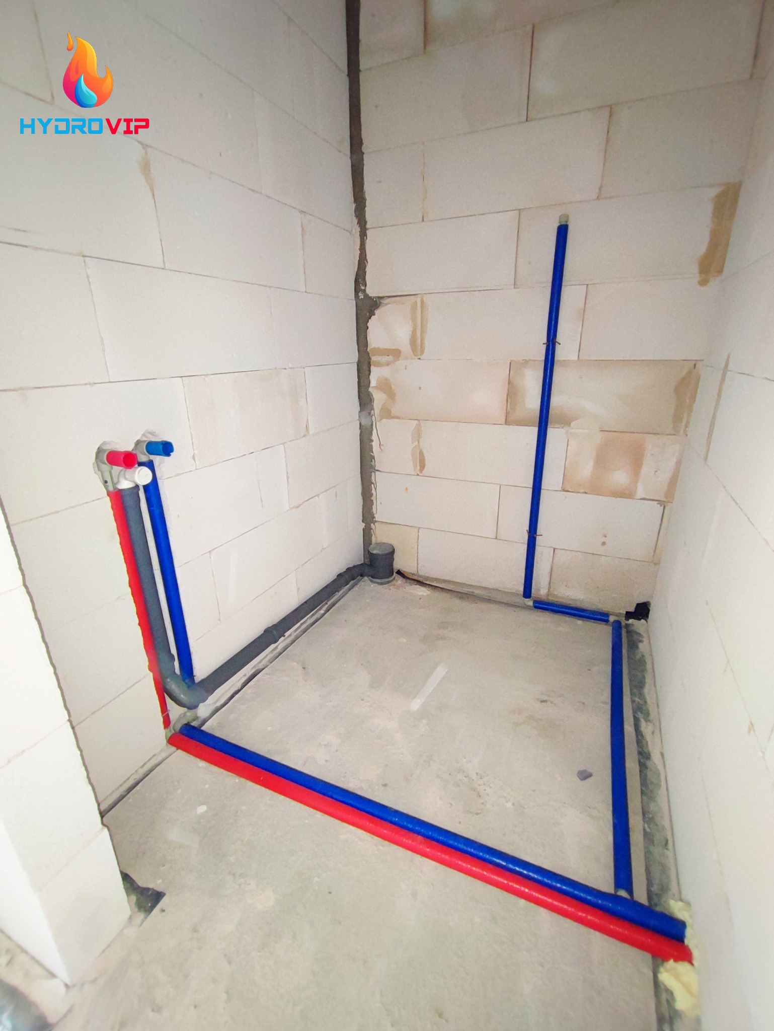 Surowy stan łazienki w trakcie prac instalacyjnych: widoczne rury wodne (czerwone i niebieskie) prowadzone wzdłuż podłogi i ścian, pionowa rura kanalizacyjna w rogu, ściany z białych bloczków.