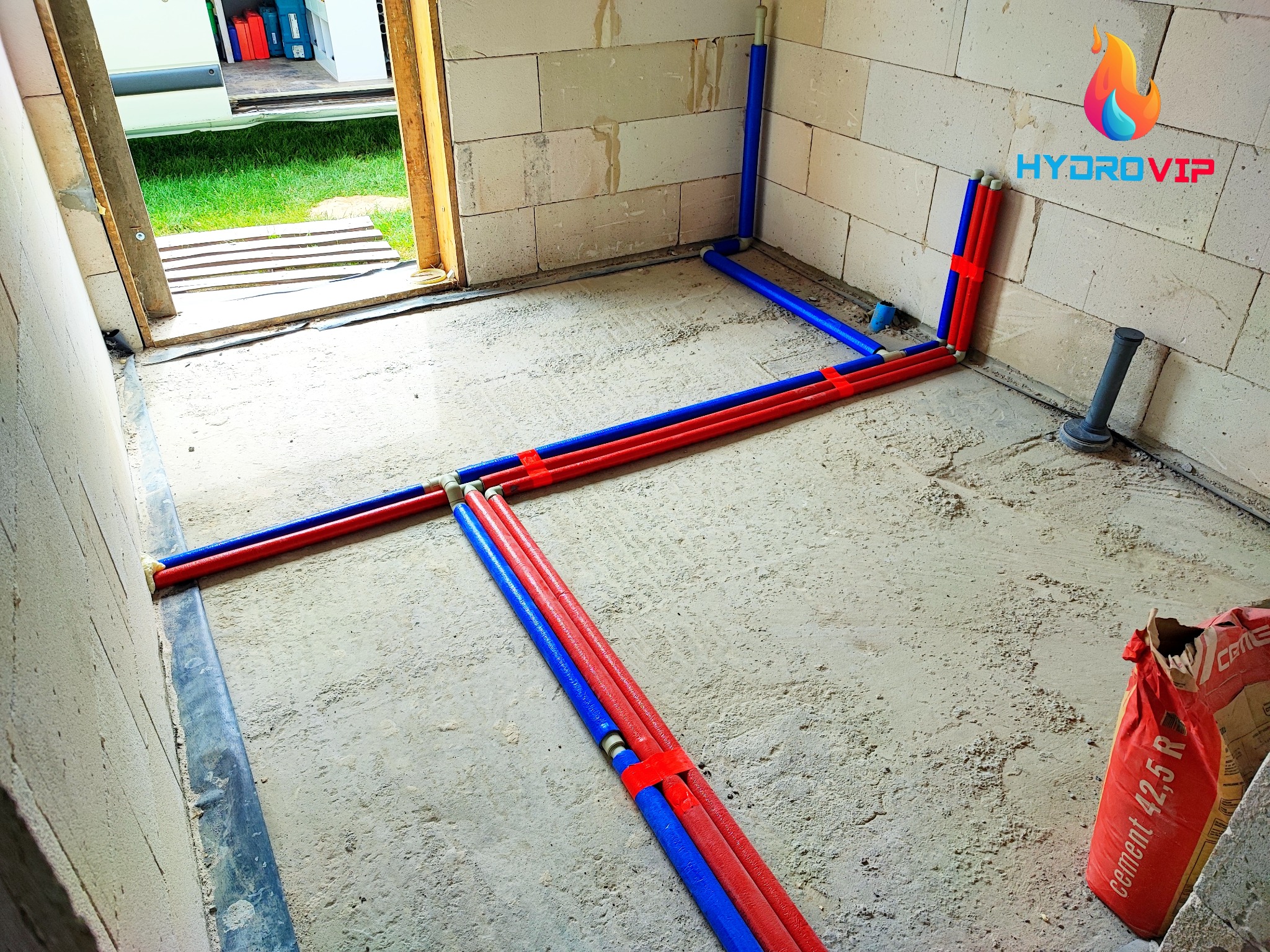 Instalacja ogrzewania podłogowego z rur PEX w kolorze czerwonym i niebieskim, mocowanych pomarańczowymi uchwytami, na surowym podkładzie betonowym w pomieszczeniu z ścianami z betonu komórkowego...