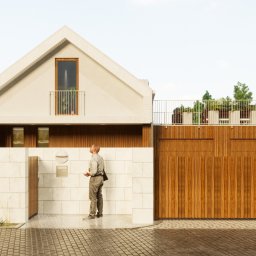 ROLKA - Pracownia Architektury - Nowoczesny dom jednorodzinny z płaskim dachem i drewnianą elewacją, mężczyzna w casualowym stroju przy skrzynce pocztowej, widoczny fragment brukowanego podjazdu i roślinność.